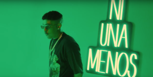 Bad Bunny wearing sunglasses & a chain in front of a green background and neon sign.