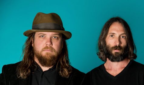 two men, one in hat, stand in front of blue background