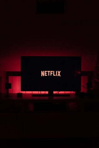 The red and white Netflix logo lights up a black flat screen tv screen in a dark room with red back lighting.