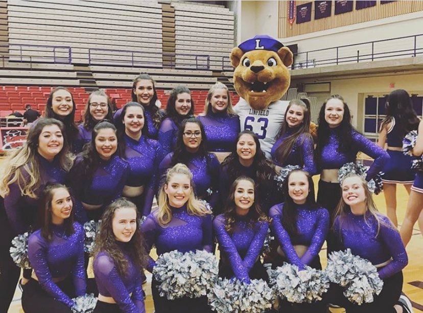 Photo+of+the+ladies+of+the+Linfield+Dance+Team+in+purple+uniforms+with+silver+pompoms+with+Mack+the+Wildcat+in+the+Linfield+gym.